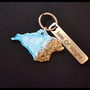 May include: A keychain featuring a miniature Isle of Wight map design. The map is a combination of blue resin representing the sea and a textured brown section. A metal tag reads "Isle Of Wight".