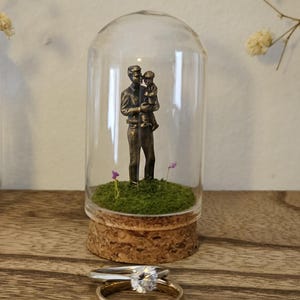 May include: A glass cloche with a cork base containing a bronze-toned figurine of an adult holding a child standing on green moss with two tiny purple flowers, accompanied by a silver-toned ring with a clear stone and a gold-toned band.