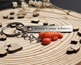 May include: A silver keychain with a lobster charm and the phrase "World's your lobster" engraved on a metal tag. The orange lobster charm is attached to a silver chain and key ring. Small wooden hearts are scattered around the keychain on a wooden surface.