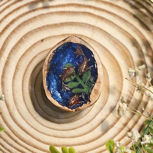 May include: A handcrafted pendant made from a walnut shell, filled with blue resin, copper-coloured accents, and a sprig of green leaves. The pendant is displayed on a wooden surface, surrounded by greenery and small white flowers.