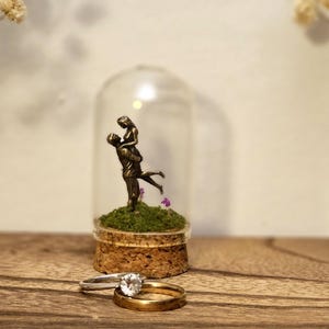 以下が含まれることがあります： 緑の苔の上に、抱き合うカップルのミニチュア像が入ったガラスドーム。ダイヤモンド付きの指輪と金の指輪が2つ前にあります。ガラスの花瓶に入ったドライフラワーがドームの両側にあります。