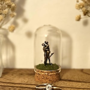 Könnte beinhalten: Eine Glaskuppel mit einer Miniatur-Bronzeskulptur eines sich umarmenden Paares, auf einem Korksockel mit grünem Moos. Zwei Ringe, einer mit einem Diamanten, liegen auf einer Holzoberfläche, was Liebe und Engagement suggeriert.