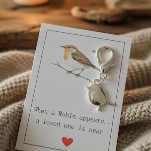 Puede incluir: Un dije de pájaro plateado cuelga de una tarjeta con una ilustración de petirrojo y el texto "When a Robin appears.. a loved one is near". Una vela encendida y una manta de punto beige están en el fondo.