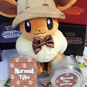 May include: A brown and tan Eevee plush toy wearing a brown hat with a small brown Eevee plush toy on top. The Eevee plush toy is wearing a brown bow tie. The Eevee plush toy is sitting on a table with a clear plastic container of wax melts and a metal tin of wax melts. The wax melts are labeled "Normal Type".