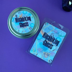 Puede incluir: Un recipiente de pl&aacute;stico transparente con un dise&ntilde;o azul y dorado. El recipiente tiene el texto "Hitchhiking Ghosts" y "Funeral Flowers, Graveyard Dirt and 090 Happy Haunts" impreso en &eacute;l. El recipiente est&aacute; sentado sobre una superficie morada.