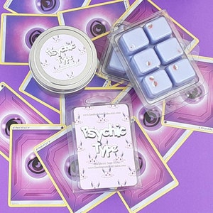 May include: Six lavender-colored wax melts in a clear plastic tray. The wax melts are decorated with small red and white sprinkles. The tray is on top of a purple background with several Pokemon cards. The cards have a purple background with a black eye in the center. The cards have the word "ENERGY" printed on them. There is a small white label on the tray that says "Psychic Type" with a website address.
