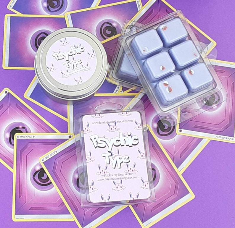 May include: Six lavender-colored wax melts in a clear plastic tray. The wax melts are decorated with small red and white sprinkles. The tray is on top of a purple background with several Pokemon cards. The cards have a purple background with a black eye in the center. The cards have the word "ENERGY" printed on them. There is a small white label on the tray that says "Psychic Type" with a website address.