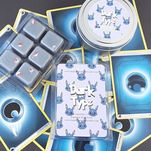 May include: Dark Type scented wax melts in a clear plastic container with six individual wax melts. The wax melts are gray with a red accent. The wax melts are surrounded by Pokemon trading cards with a blue and yellow design. The cards have the text "Dark Type" and "Energy".