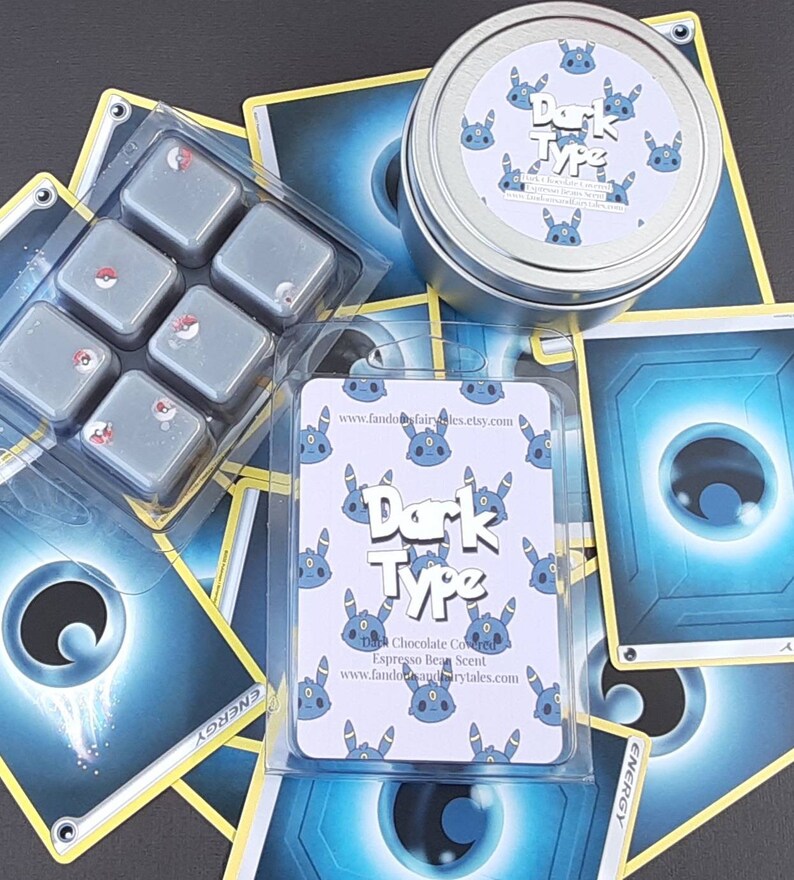 May include: Dark Type scented wax melts in a clear plastic container with six individual wax melts. The wax melts are gray with a red accent. The wax melts are surrounded by Pokemon trading cards with a blue and yellow design. The cards have the text "Dark Type" and "Energy".