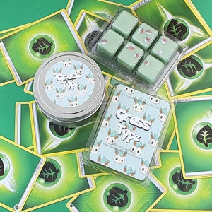 May include: A set of six green wax melts in a clear plastic container. The wax melts are shaped like squares and have a white design on them. The container is sitting on top of a stack of green and yellow Pokemon cards. The cards have a green leaf design on them and the word "ENERGY" printed on them. The wax melts are labeled "Grass Type" and have a website address printed on them.