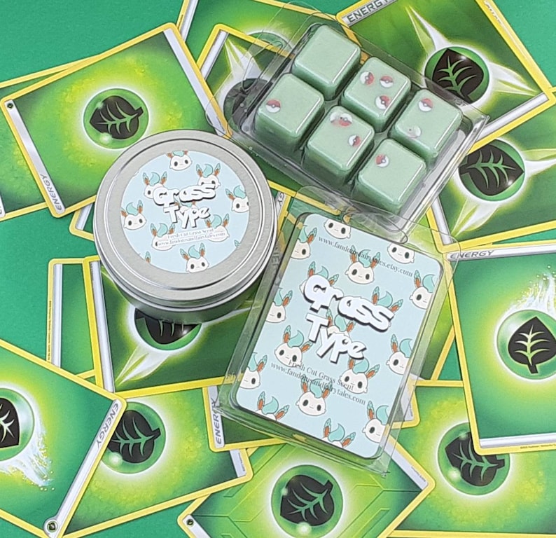 May include: A set of six green wax melts in a clear plastic container. The wax melts are shaped like squares and have a white design on them. The container is sitting on top of a stack of green and yellow Pokemon cards. The cards have a green leaf design on them and the word "ENERGY" printed on them. The wax melts are labeled "Grass Type" and have a website address printed on them.
