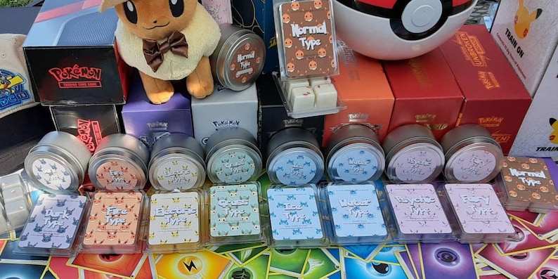 May include: A collection of 10 small, round, silver tins with labels featuring different Pokemon types. The labels are colorful and feature the type name in white text. The tins are arranged in two rows of five. The tins are sitting on a table with a variety of Pokemon cards scattered around them.