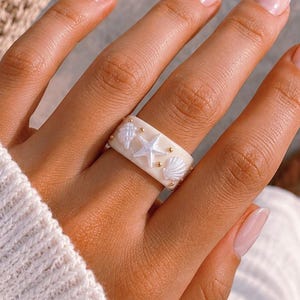 May include: A wide, cream-coloured band ring featuring a raised white starfish and seashell embellishments. The ring is decorated with small, metallic gold-toned beads placed between the shell motifs, creating a textured, nautical-inspired design.