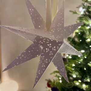 May include: A lavender paper star decoration with a detailed, perforated design. The star has eight points and is hung by a cream ribbon. A Christmas tree with fairy lights is in the background, suggesting a festive holiday theme.