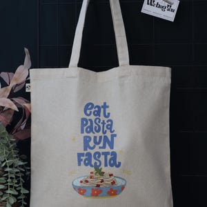 Peut inclure: Sac fourre-tout en toile beige avec le texte bleu "Eat Pasta Run Fasta". En dessous, un dessin d'un bol de pâtes. Le sac a de longues anses de couleur crème.