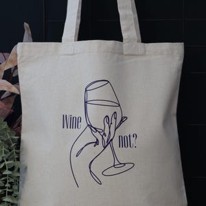 Op de afbeelding: Beige canvas draagtas met een donkerblauwe lijntekening van een wijnglas dat door een hand wordt vastgehouden. De woorden "Wine not?" zijn in donkerblauw op de tas gedrukt.