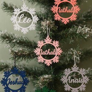 Op de afbeelding: Sneeuwvlokvormige kerstversieringen in wit, koraal, roze, blauw, groen en grijs, elk met een naam uitgesneden in het midden. De ornamenten hangen aan een kerstboom. Namen zijn Léo, Nathali, Joshua, Anais en Hugo.