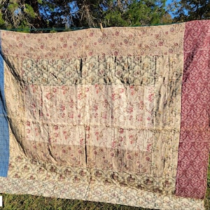 May include: A patchwork quilt, approximately 85 x 70 inches, hangs on a clothesline. The quilt features floral patterns in cream, beige, and burgundy, with a blue border and a burgundy patterned edge. The quilt is made of multiple fabric panels.