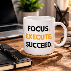 May include: White ceramic mug with the words "FOCUS. EXECUTE. SUCCEED." in black and orange lettering. The mug sits on a cork coaster on a wooden desk, next to a notebook and pen. A laptop and plant are in the background.