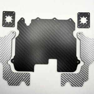 May include: A collection of carbon fibre components, including a large central piece, two smaller pieces with a silver and black weave, and two square pieces with a star-shaped cutout. The pieces are arranged on a white surface.