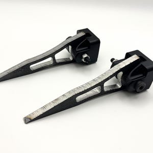 May include: Two black and silver metal brackets with a triangular design. The brackets have a textured, silver-coloured finish on the angled arms and a black base with a screw. The items are displayed on a white background.