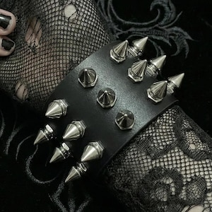 Może przedstawiać: Czarna skórzana bransoletka ze srebrnymi stożkowymi ćwiekami. Bransoletka to odważny dodatek, z rzędem dużych, spiczastych ćwieków i mniejszych, zaokrąglonych ćwieków. Projekt osadzony jest na tle czarnej koronki i tkaniny, tworząc gotycką estetykę.