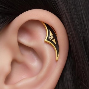 Puede incluir: Un pendiente de oreja dorado con incrustaciones de esmalte negro. El pendiente presenta un diseño estilizado con un borde dorado y un símbolo de trébol de tres hojas. La joya se lleva en la oreja.