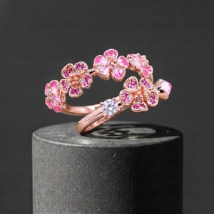 Può includere: Un anello in oro rosa con un design floreale con petali in smalto rosa e piccole pietre preziose rosa. L'anello presenta anche una gemma rotonda e trasparente. L'anello è esposto su una superficie grigio scuro.