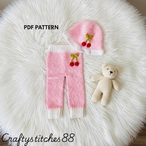 May include: A crochet PDF pattern set featuring pink and white pants and a matching hat, both decorated with small red cherry appliques with green stems. A small, light beige crocheted teddy bear is positioned next to the clothing items.