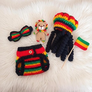 May include: A crochet set featuring a nappy cover, a bow tie, a small lion plush, and a hat with black yarn dreadlocks. The items are crafted in red, yellow, green, and black yarn. The hat is topped with a striped cap, and a small flag is included.