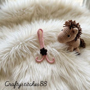 Peut inclure: Une lettre I crochet&eacute;e rose clair avec une petite fleur marron fonc&eacute;, plac&eacute;e &agrave; c&ocirc;t&eacute; d'un petit cheval en crochet marron clair avec une crini&egrave;re et une queue marron fonc&eacute;. Le texte Craftystitches88 est visible au bas de l'image.
