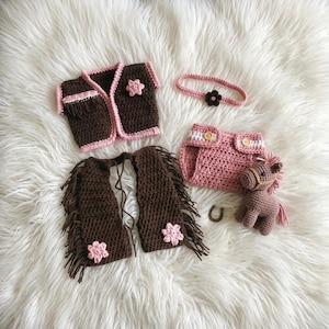 Peut inclure: Un ensemble au crochet comprenant un gilet marron fonc&eacute; avec bordure rose et une fleur, des jambi&egrave;res marron &agrave; franges avec fleurs roses, une culotte rose &agrave; deux boutons, un bandeau rose avec une fleur fonc&eacute;e et un petit cheval en crochet.