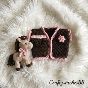 Peut inclure: Un gilet au crochet marron avec des bordures roses, une frange et une applique en forme d'&eacute;toile rose, accompagn&eacute; d'un petit cheval au crochet beige avec une crini&egrave;re, une queue et des sabots marron fonc&eacute;, orn&eacute; d'un n&oelig;ud rose. Texte : CraftyStitches88.