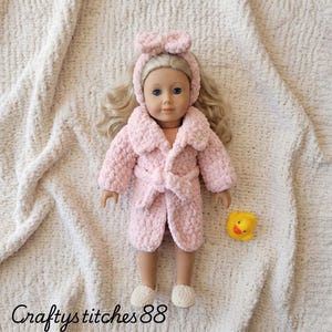 Peut inclure: Une poupée aux cheveux blonds portant un peignoir en crochet rose clair avec un bandeau assorti et des chaussons blancs. La poupée est accompagnée d'un petit canard en caoutchouc jaune. Le texte Craftystitches88 est visible en bas à gauche.