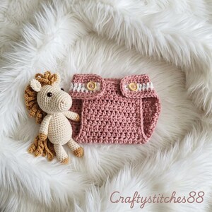 Peut inclure: Un jouet cheval crochet&eacute; de couleur cr&egrave;me avec une crini&egrave;re et une queue beige, plac&eacute; &agrave; c&ocirc;t&eacute; d'une culotte de protection crochet&eacute;e rose poudr&eacute; avec une bande horizontale blanche et deux boutons en bois clair. Le texte CraftyStitches88 est visible.