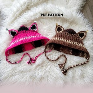 May include: Two crochet hats with animal ears and braided ties. One hat is bright pink with black stripes and white accents, the other is brown with dark brown stripes and cream accents. Both feature small metallic charms. Text reads PDF PATTERN.