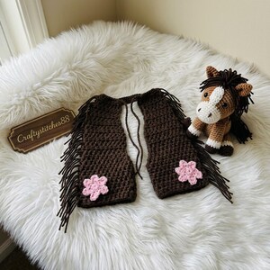 Peut inclure: Un gilet au crochet marron fonc&eacute; avec de longues franges sur les bords et une fleur au crochet rose clair sur chaque panneau avant. &Agrave; c&ocirc;t&eacute; du gilet se trouve un petit cheval en crochet avec une crini&egrave;re marron fonc&eacute;, une liste blanche et des sabots blancs.