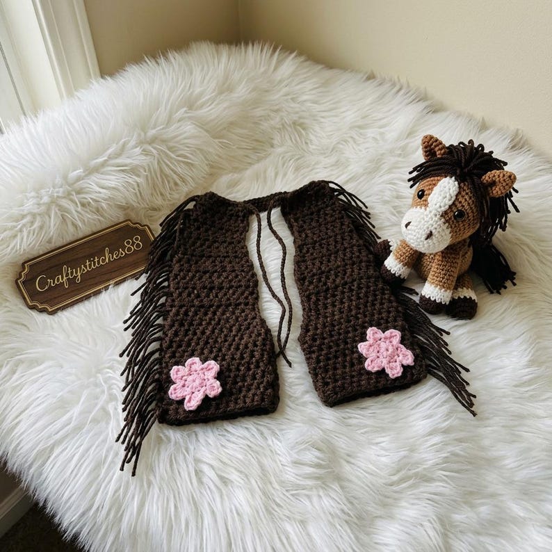 Peut inclure: Un gilet au crochet marron fonc&eacute; avec de longues franges sur les bords et une fleur au crochet rose clair sur chaque panneau avant. &Agrave; c&ocirc;t&eacute; du gilet se trouve un petit cheval en crochet avec une crini&egrave;re marron fonc&eacute;, une liste blanche et des sabots blancs.
