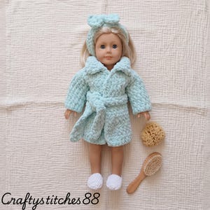 Peut inclure: Une poupée aux cheveux blonds portant un peignoir en peluche texturé bleu clair, un bandeau assorti avec un nœud et des chaussons blancs. À côté de la poupée se trouvent une éponge beige naturelle et une brosse à cheveux en bois.