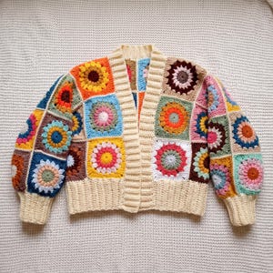 May include: A long-sleeved cardigan constructed from multicoloured crochet granny squares featuring floral motifs in shades of orange, yellow, pink, blue, and brown. The garment has a cream-coloured ribbed trim along the front opening and cuffs.
