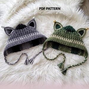 Puede incluir: Dos gorros de ganchillo con orejas de gato y lazos trenzados. Un gorro es gris y negro con cascabeles plateados en las orejas, y el otro es verde y verde oscuro con cascabeles dorados. El texto PDF PATTERN es visible en la parte superior.