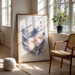 a large framed painting of a serene landscape with mountains and trees, placed on a wooden floor in a sunlit room. Next to the painting is a woven chair, and a potted plant is visible in the background.