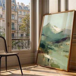 a large, framed painting of a mountainous landscape, placed on a wooden floor next to a black woven chair. The painting is leaning against a wall, and the chair is positioned in front of a window that offers a view of a cityscape.