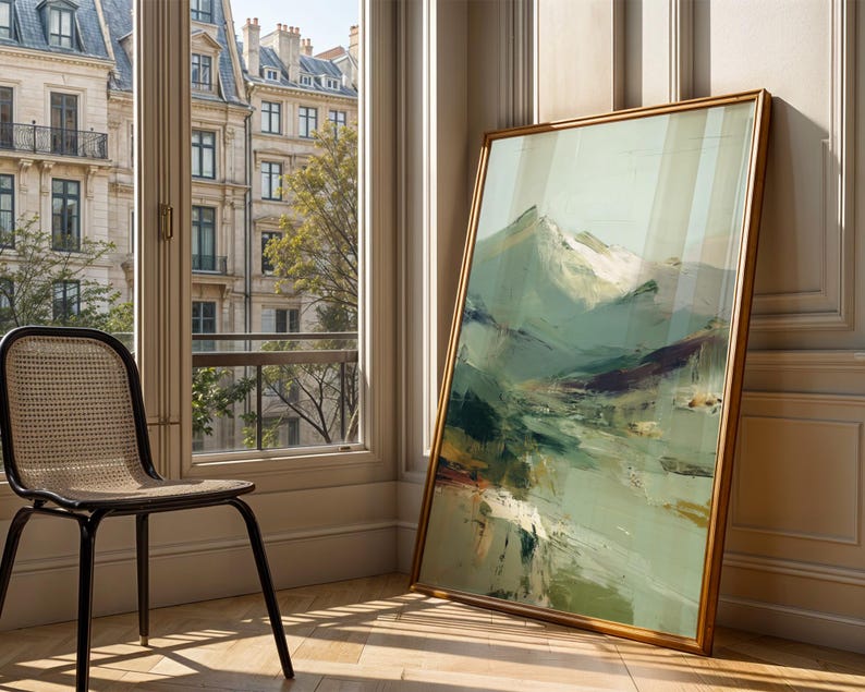 a large, framed painting of a mountainous landscape, placed on a wooden floor next to a black woven chair. The painting is leaning against a wall, and the chair is positioned in front of a window that offers a view of a cityscape.
