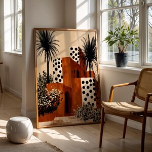 a large framed painting of a desert-like landscape with palm trees and buildings, placed in a bright and sunny room with large windows.