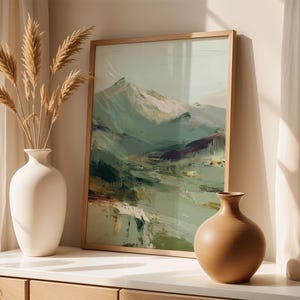 a framed landscape painting on a white shelf, accompanied by two vases. The painting depicts a mountainous scene with a mix of green, brown, and white colors. The vases are white and have a simple, elegant design. The arrangement creates a harmonious and visually appealing display.