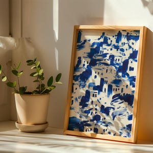 a potted plant with green leaves and a framed painting of a blue and white cityscape on a white wall.
