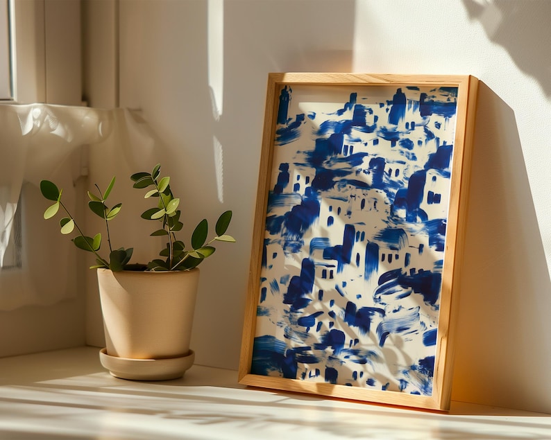 a potted plant with green leaves and a framed painting of a blue and white cityscape on a white wall.