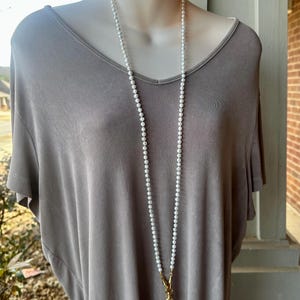 Puede incluir: Un collar largo de perlas blancas con un cierre dorado. El collar lo lleva un maniquí que lleva una camisa gris.