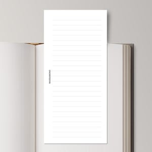 May include: A white lined notepad with the word "MASSAMONO" printed vertically on the left side is placed inside an open book. The book has a light brown cover and white pages. The background is a light grey.